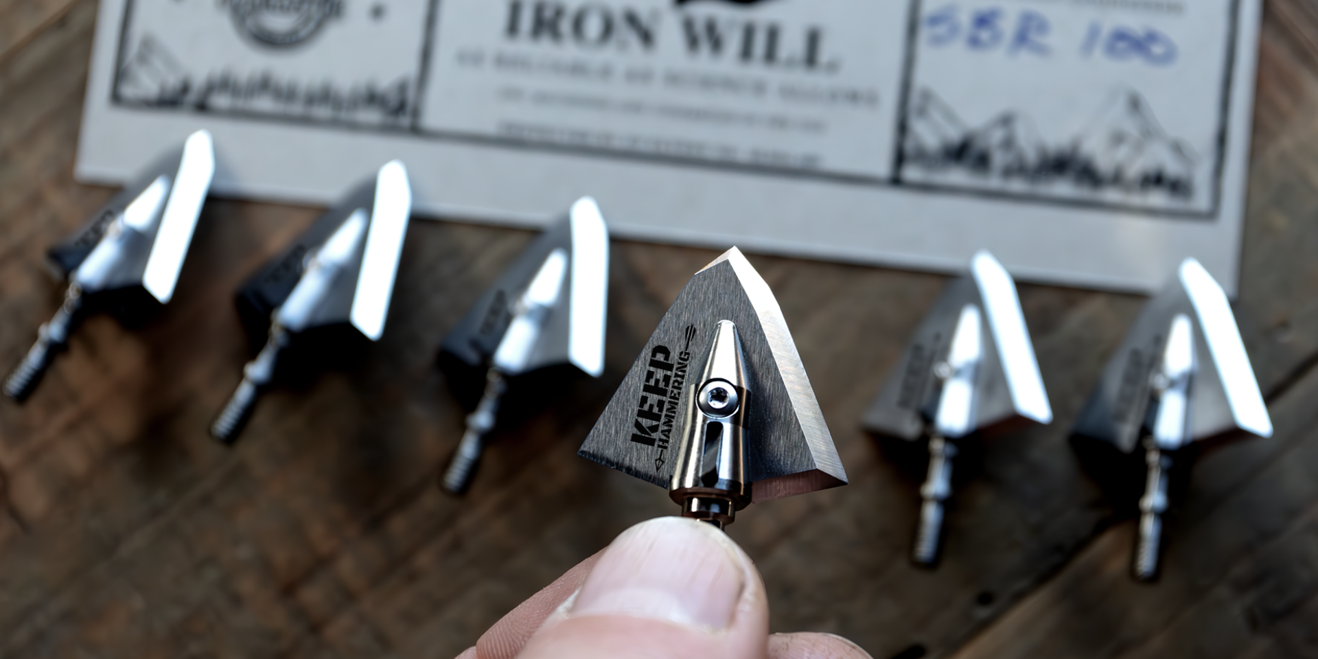 slew of broadheads on a table with piece of paper above them, with fingers holding a broadhead in the foreground that says keep hammering