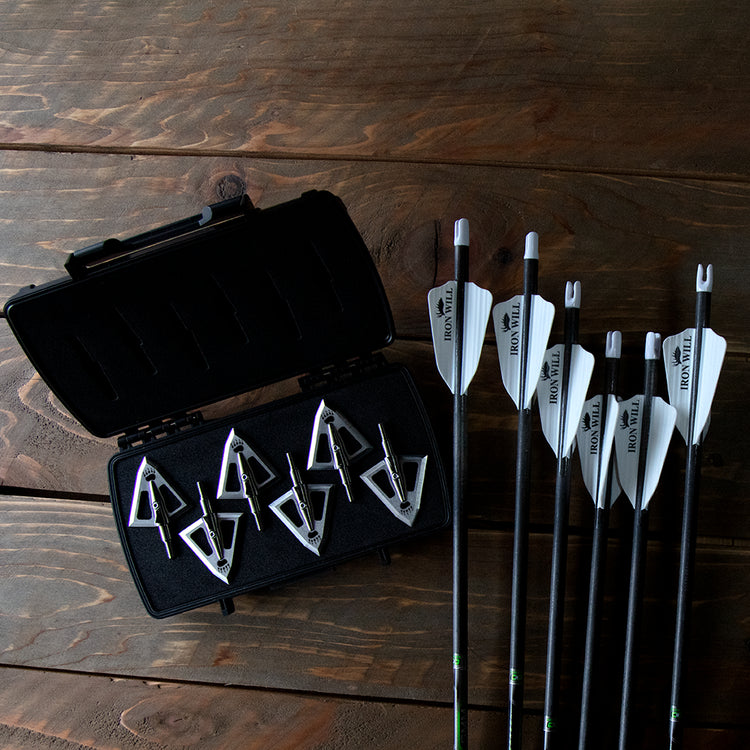 Six bear paw engraved broadheads in a case alongside arrows over a wood table