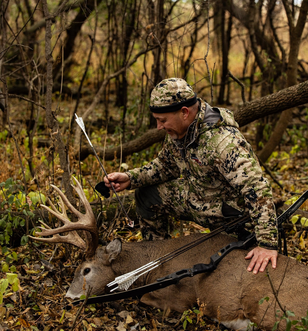 Traditional hunter in camouflage gear with a bow, arrow, and Iron Will broadhead next to a large deer in a forest setting