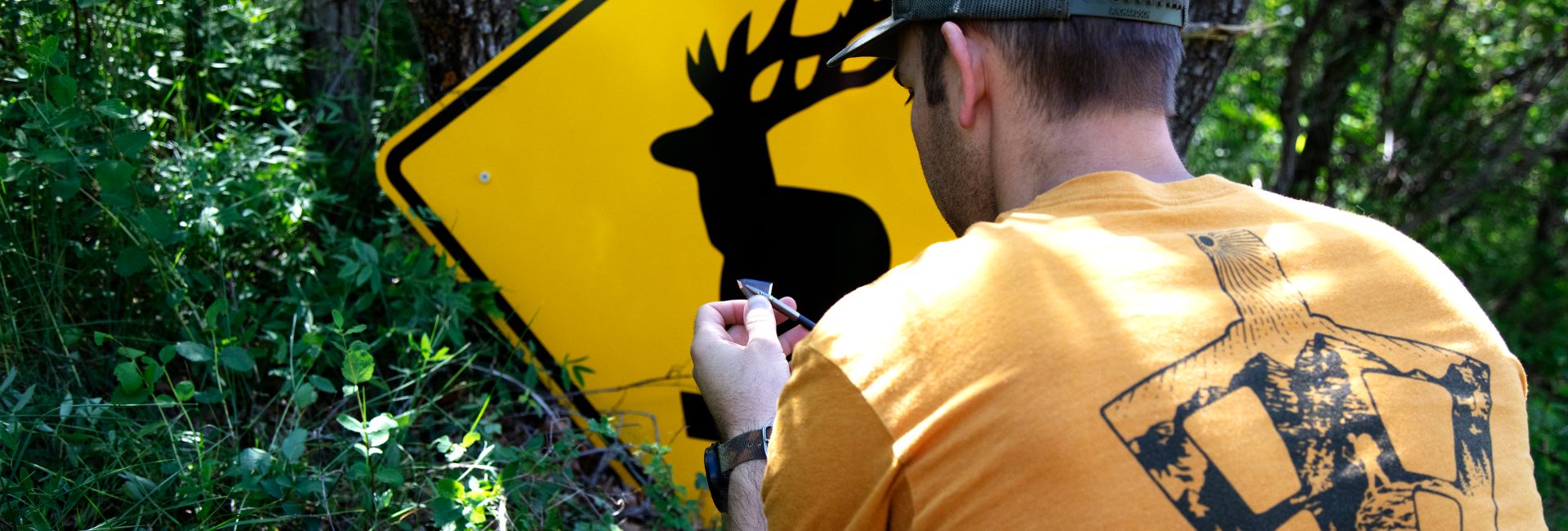 person in a yellow shirt screwing on a broadhead in front of a road sign with an elk silhouette, out in the forest.