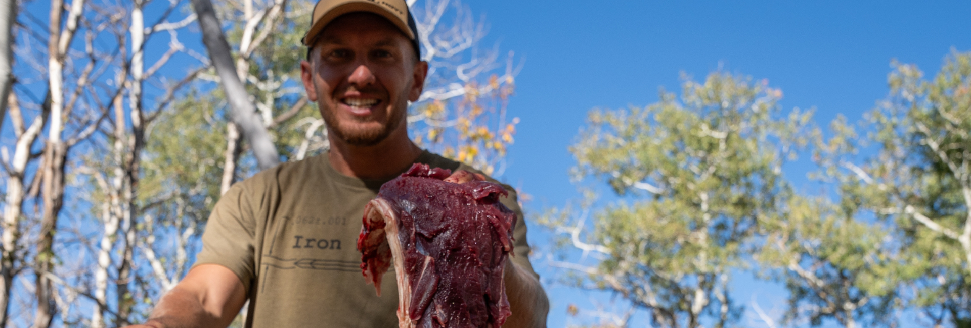 Man holding a large piece of deboned raw meat outdoors with trees and blue sky in the background