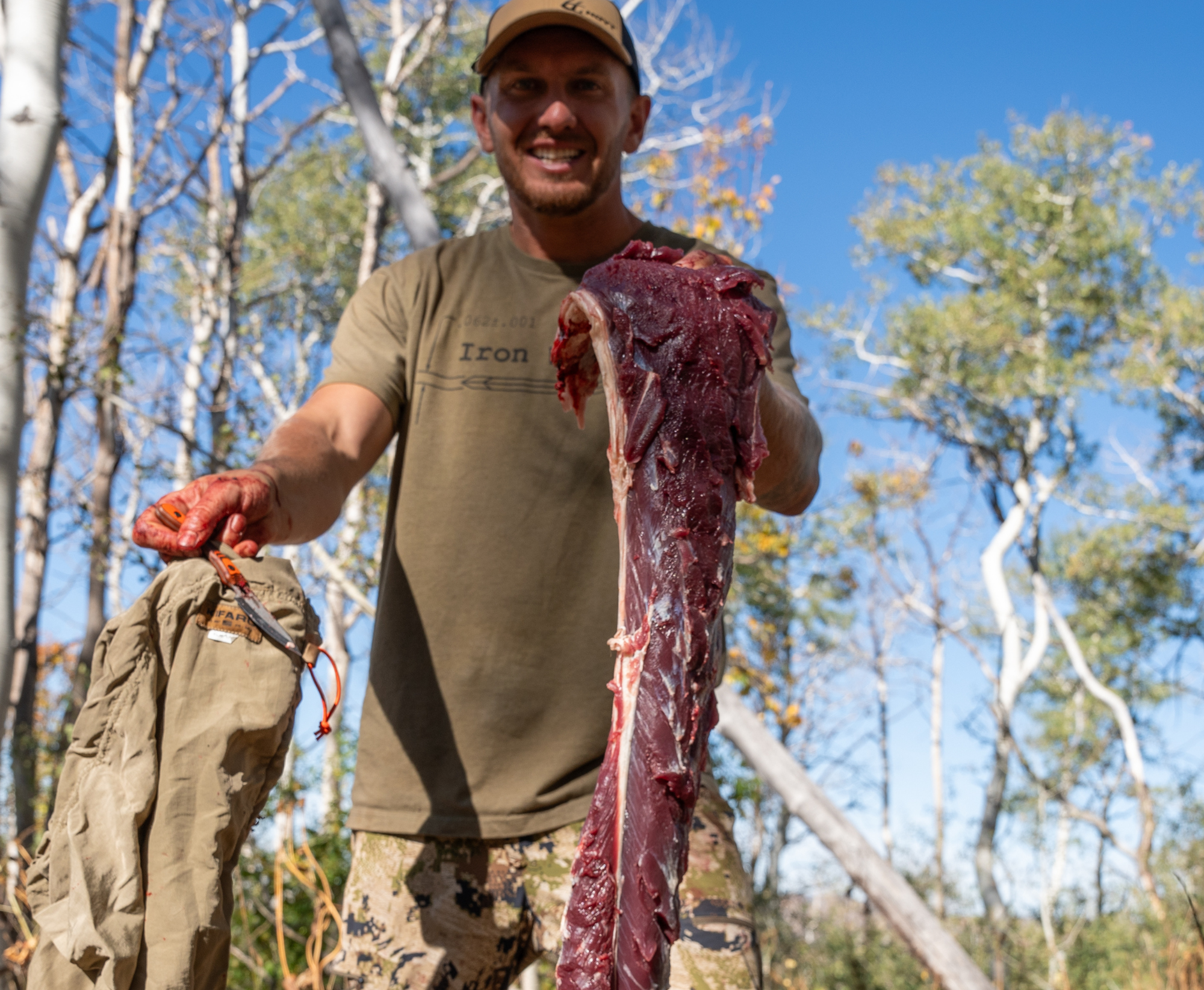 Hunter holding a large piece of deboned raw meat outdoors with trees and blue sky in the background